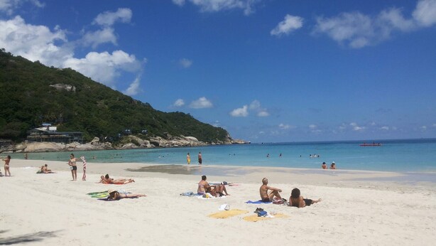 Una playa con gente tomando el sol y nadando, cielo despejado y un promontorio rocoso al fondo.