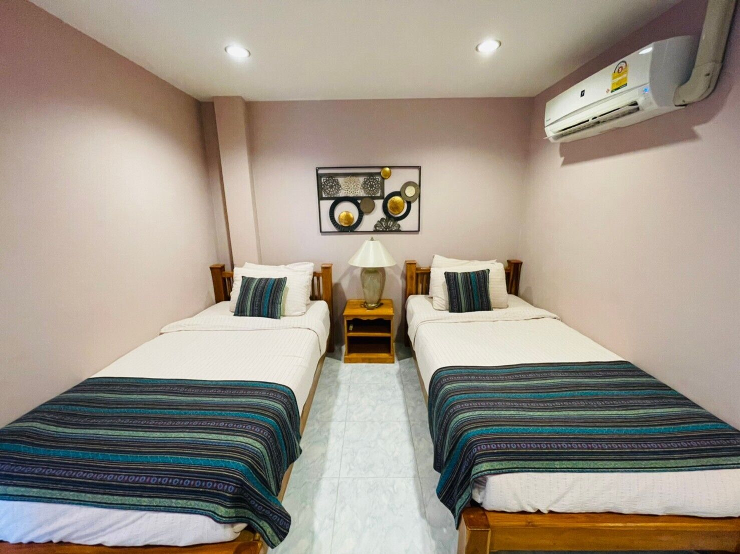 A twin room with two beds, a nightstand with a lamp, a wall-mounted air conditioner, and a decorative plate on the wall.