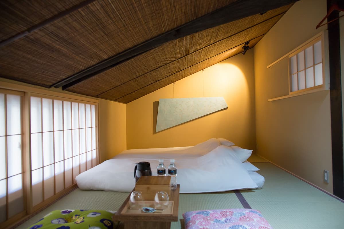 A traditional Japanese-style room with tatami flooring, a low ceiling, and sliding shoji doors. There is a bed with pillows and a small table with a kettle, cups, and a bottle.