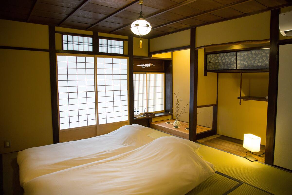 A traditional Japanese room with tatami flooring, a bed with white bedding, a hanging lantern, and sliding shoji doors.