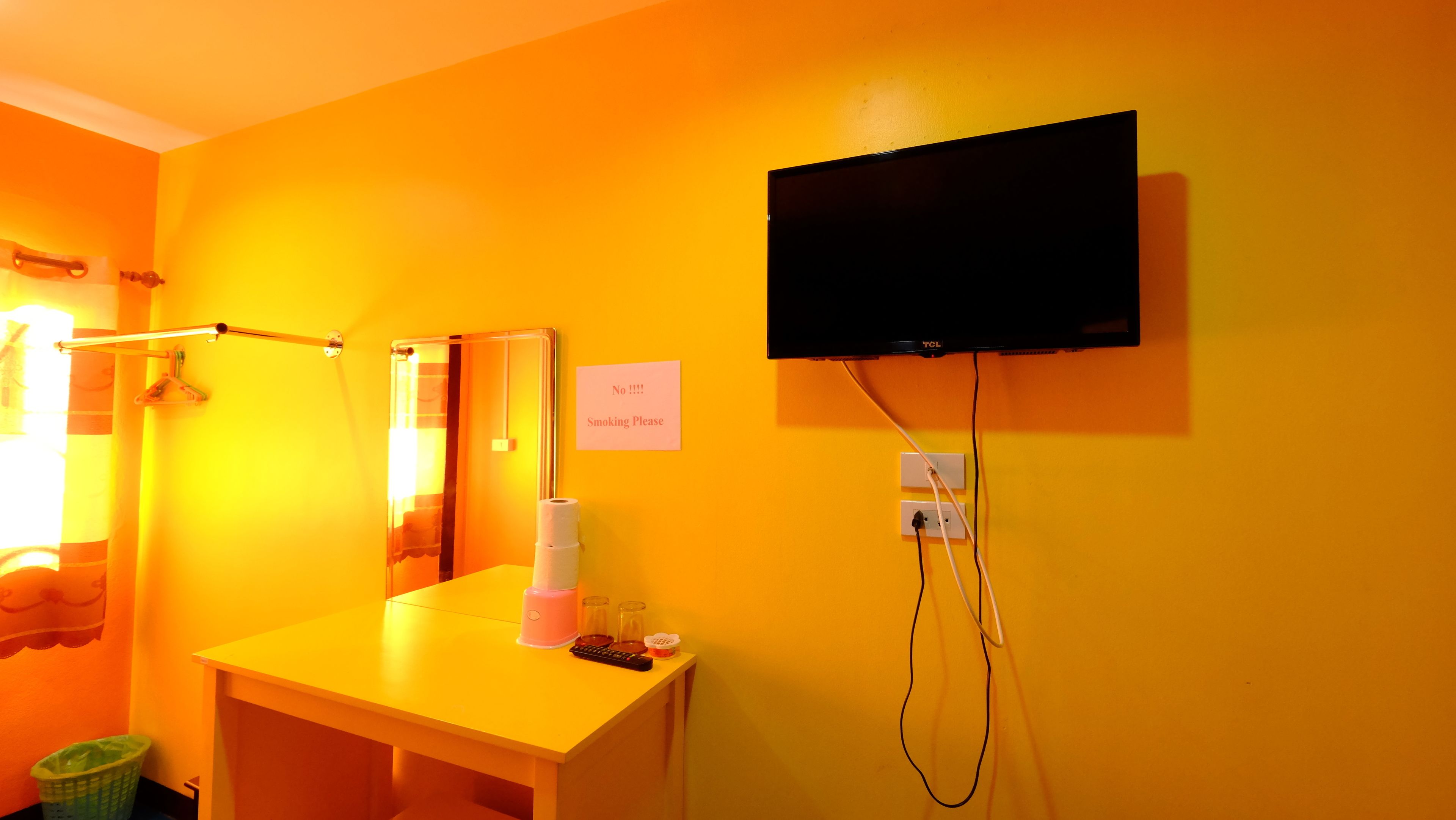 A room with a yellow wall, a black TV mounted on the wall, a towel rack, a sign prohibiting smoking, and a trash bin.