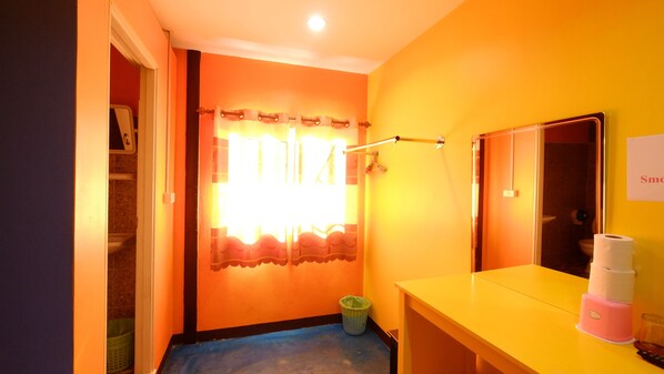 Une salle de bain avec un mur jaune, une fenêtre avec des rideaux, un miroir, un lavabo avec un distributeur de savon rose et une poubelle.