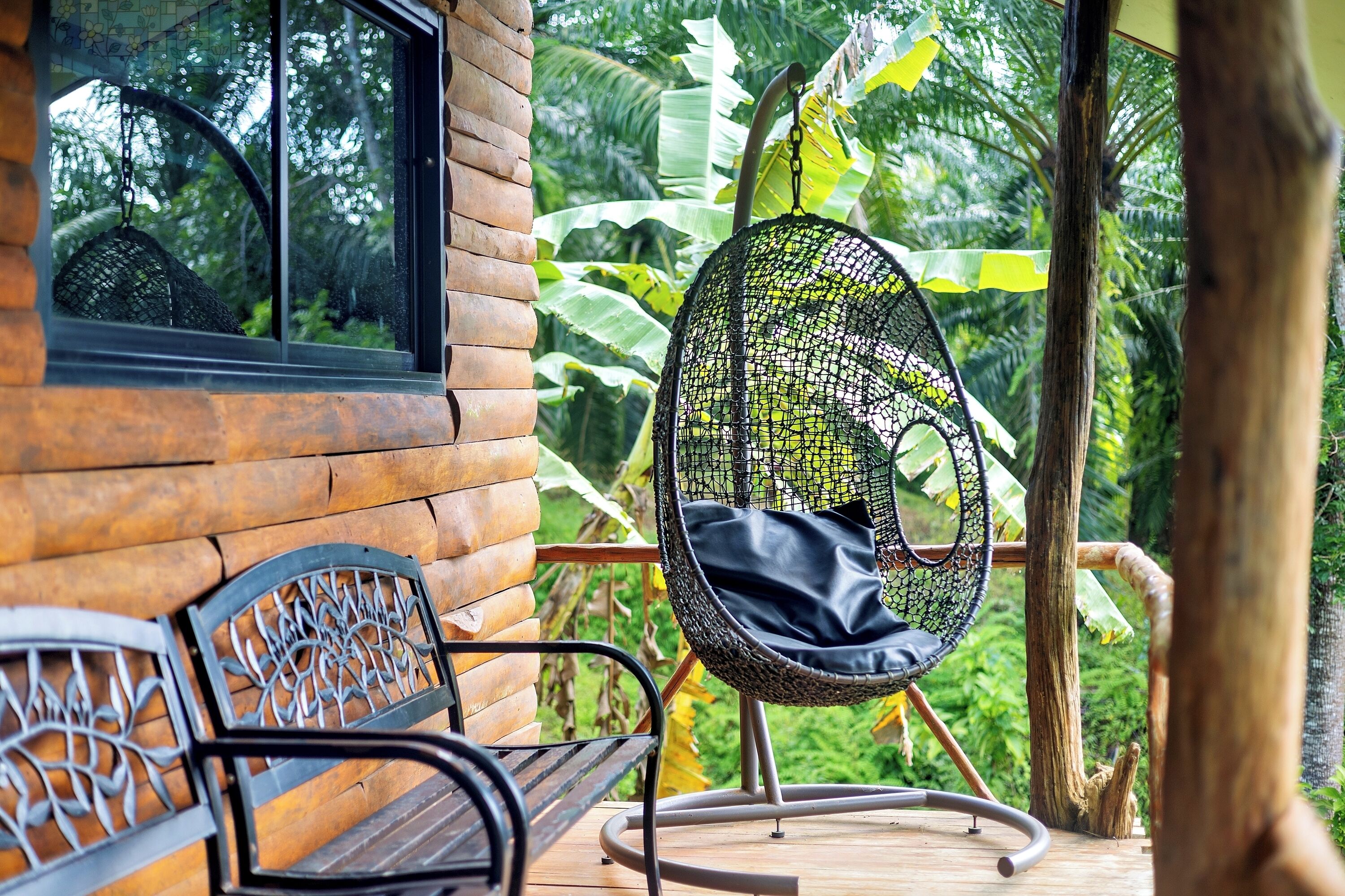 A wooden bench and a hanging chair with a black cushion on a wooden deck, surrounded by lush greenery.