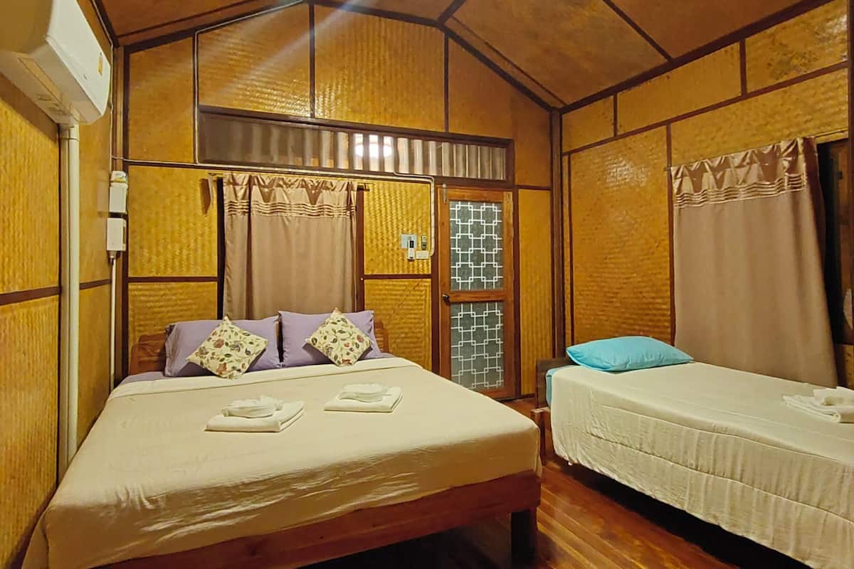 Una habitación con una cama, una cama adicional, suelo de madera, ventilador de techo y una ventana con cortinas.
