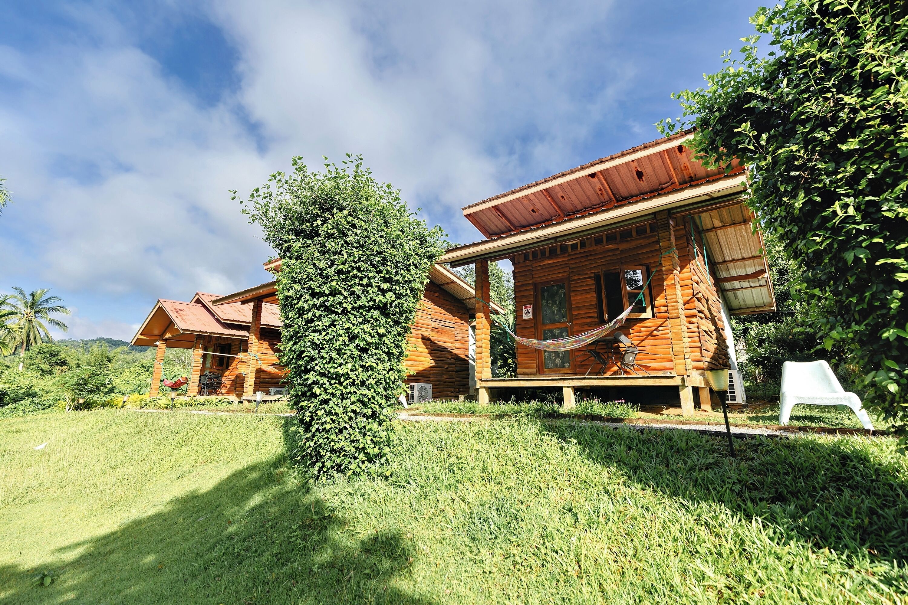 A wooden cabin with a porch, surrounded by lush greenery and a clear sky.