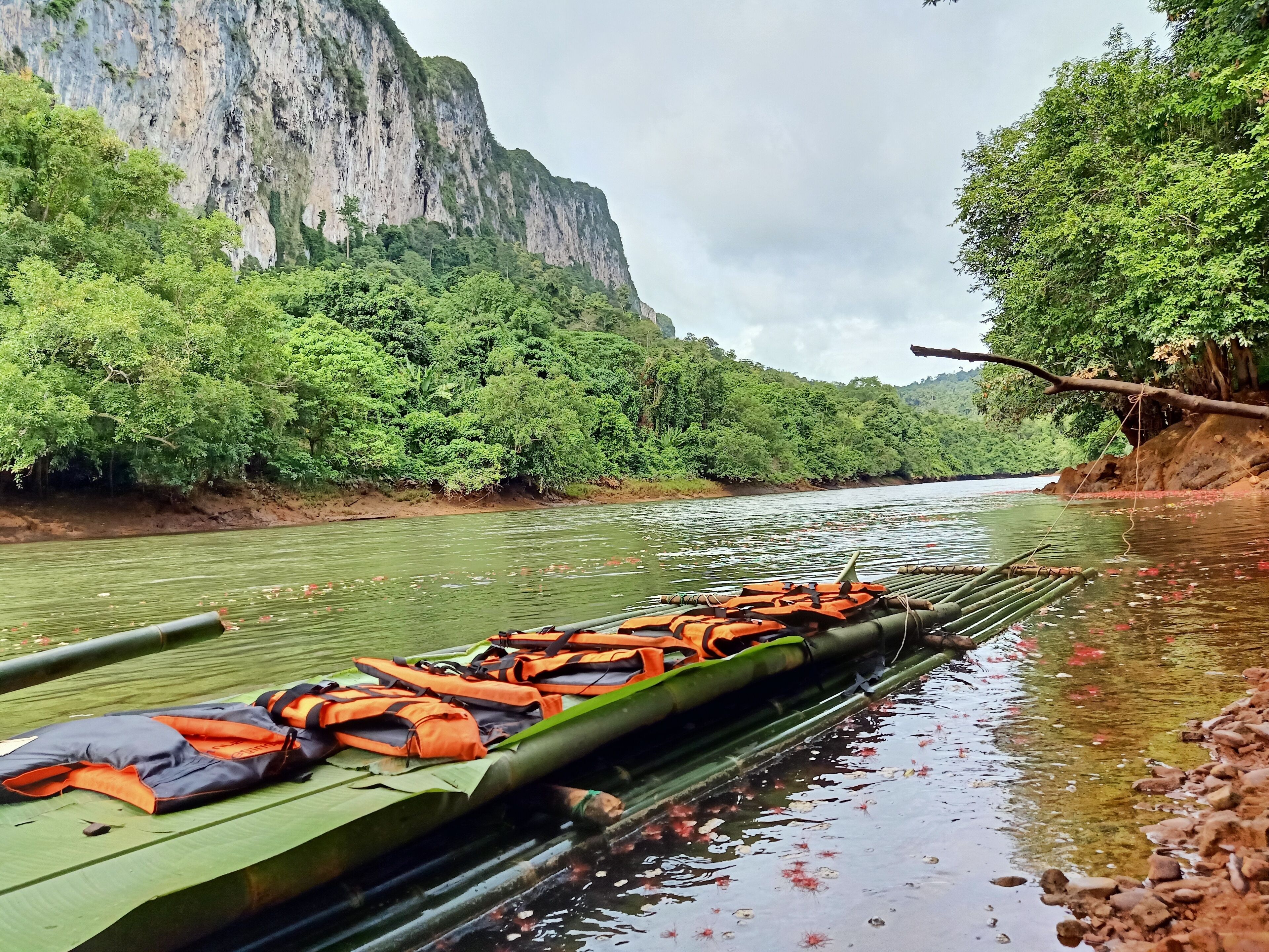 A river with a long boat carrying orange life jackets, surrounded by dense green foliage and a rocky cliff.