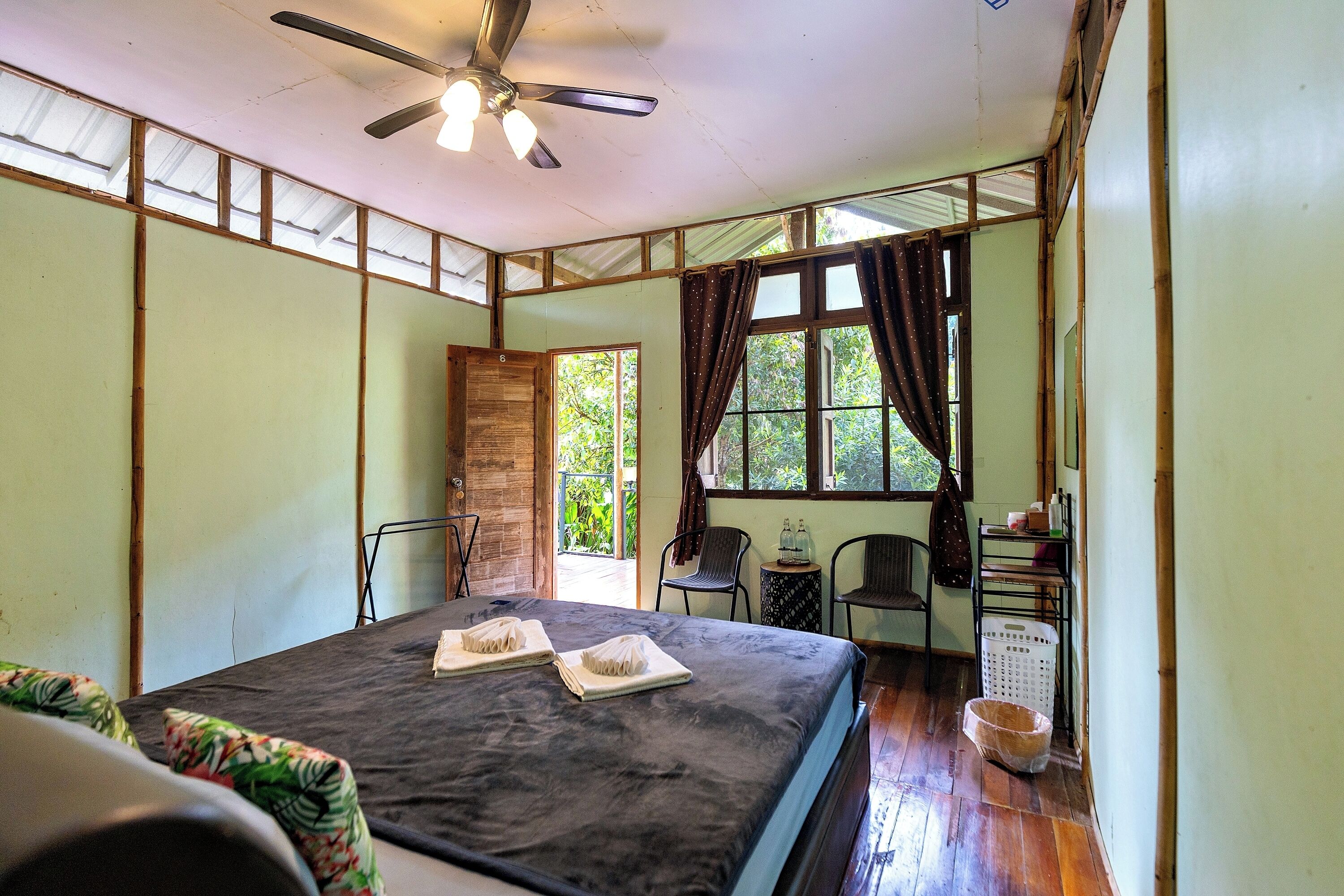 A room with a bed, ceiling fan, window with curtains, chairs, and a small table with items.