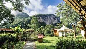 A rustic lodge with a thatched roof, surrounded by lush greenery and a mountain backdrop. - Bansuanphutarn (Ban Ta Khun)