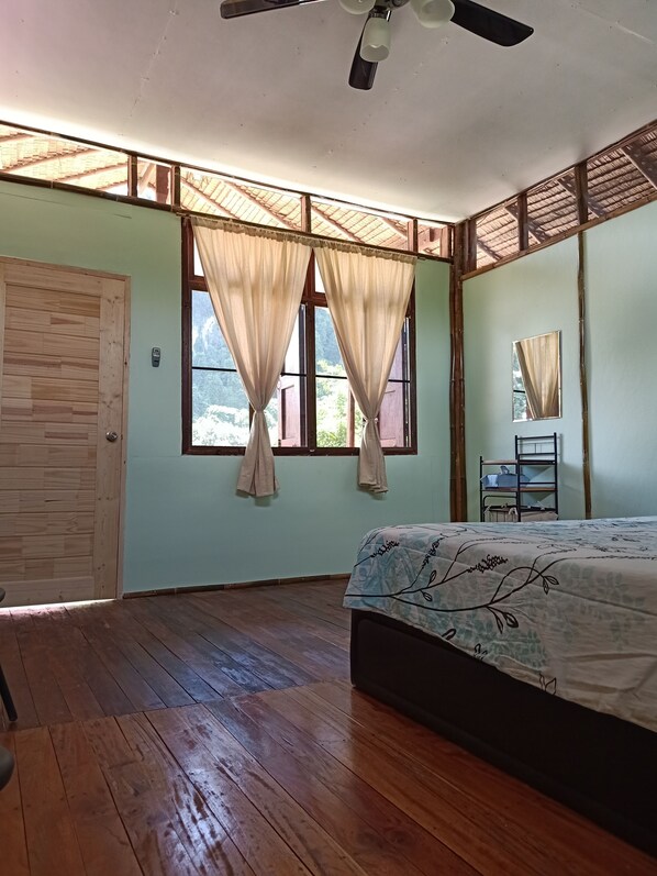 Une chambre avec un lit, un plancher en bois, un ventilateur de plafond et une fenêtre avec des rideaux.