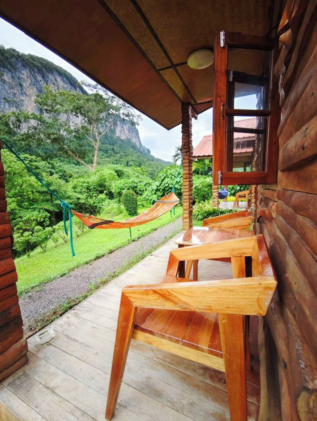 Une cabane en bois dotée d’une véranda, d’un hamac et offrant une vue sur un paysage verdoyant luxuriant, avec une montagne en arrière-plan.