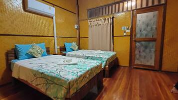 A room with two beds, wooden flooring, an air conditioning unit, a window with curtains, and a door with frosted glass panels.