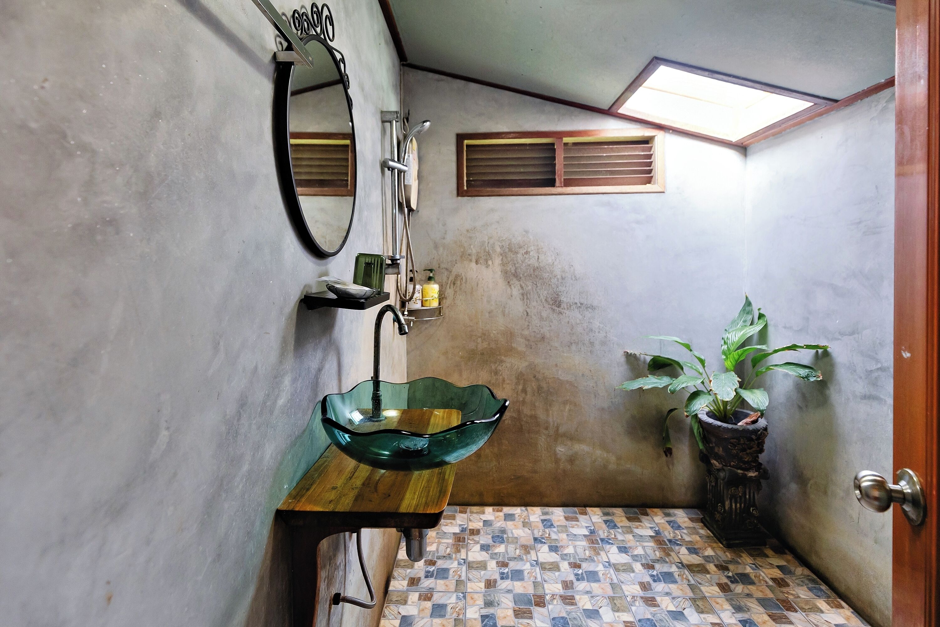 Un bany amb un lavabo de vidre, un mirall, una planta en test i un terra solcat.
