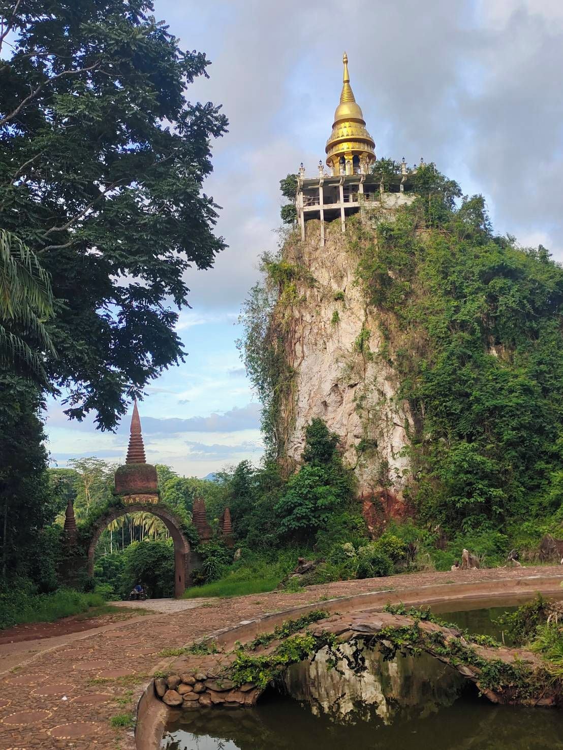 A temple on a cliff with a golden spire, surrounded by lush greenery and a small bridge over a water body.