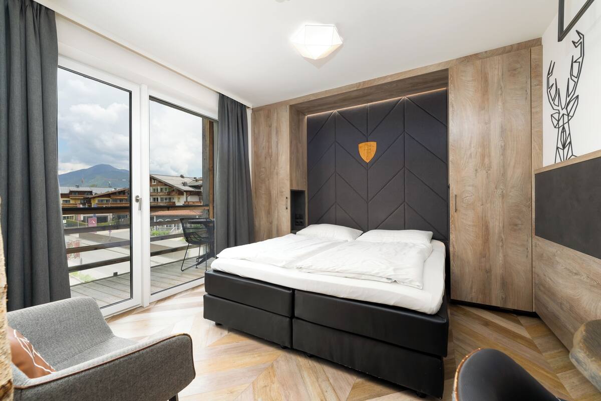 A modern bedroom with a large bed, a sliding glass door to a balcony, and a view of buildings and mountains.