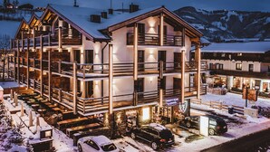 A multi-story hotel with a restaurant on the ground floor, outdoor seating, and a view of snow-covered mountains. - 24 by AvenidA - Mountain Hotel (Kaprun)