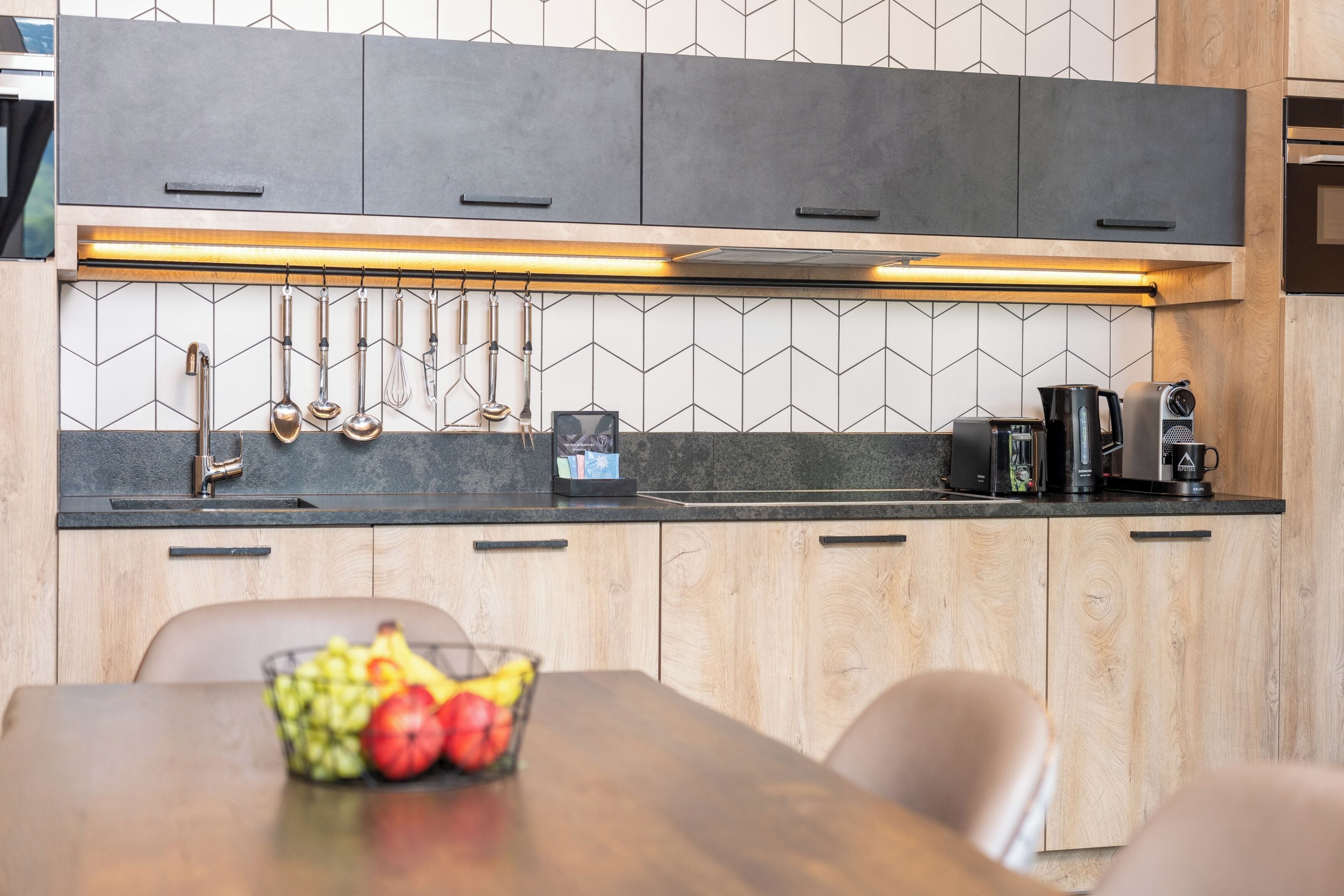 A modern kitchen with dark cabinets, a black countertop, and a wooden dining table with a fruit bowl.