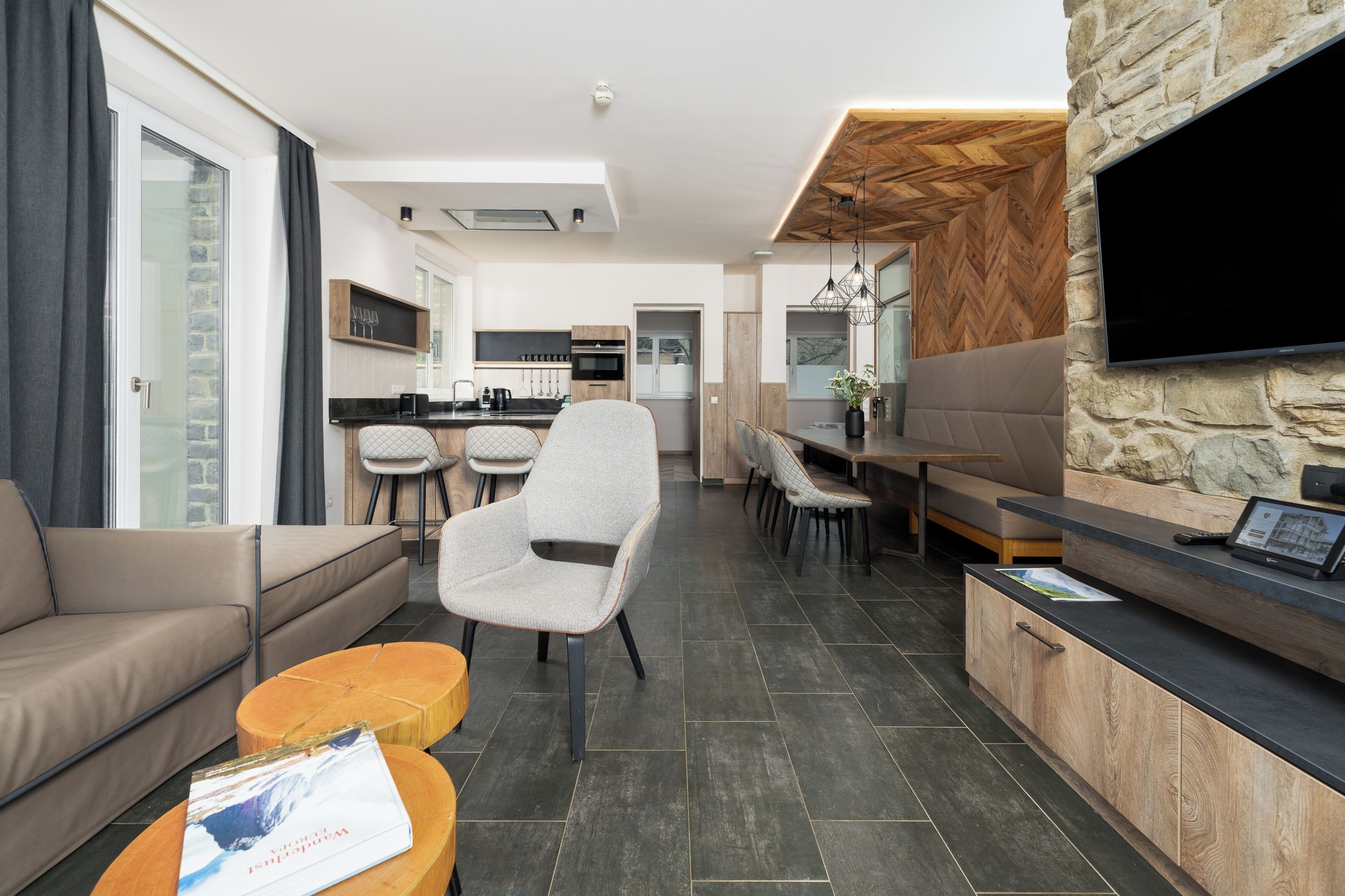 A modern living room with a sofa, armchairs, a coffee table, and a dining area with a TV mounted on a stone wall.