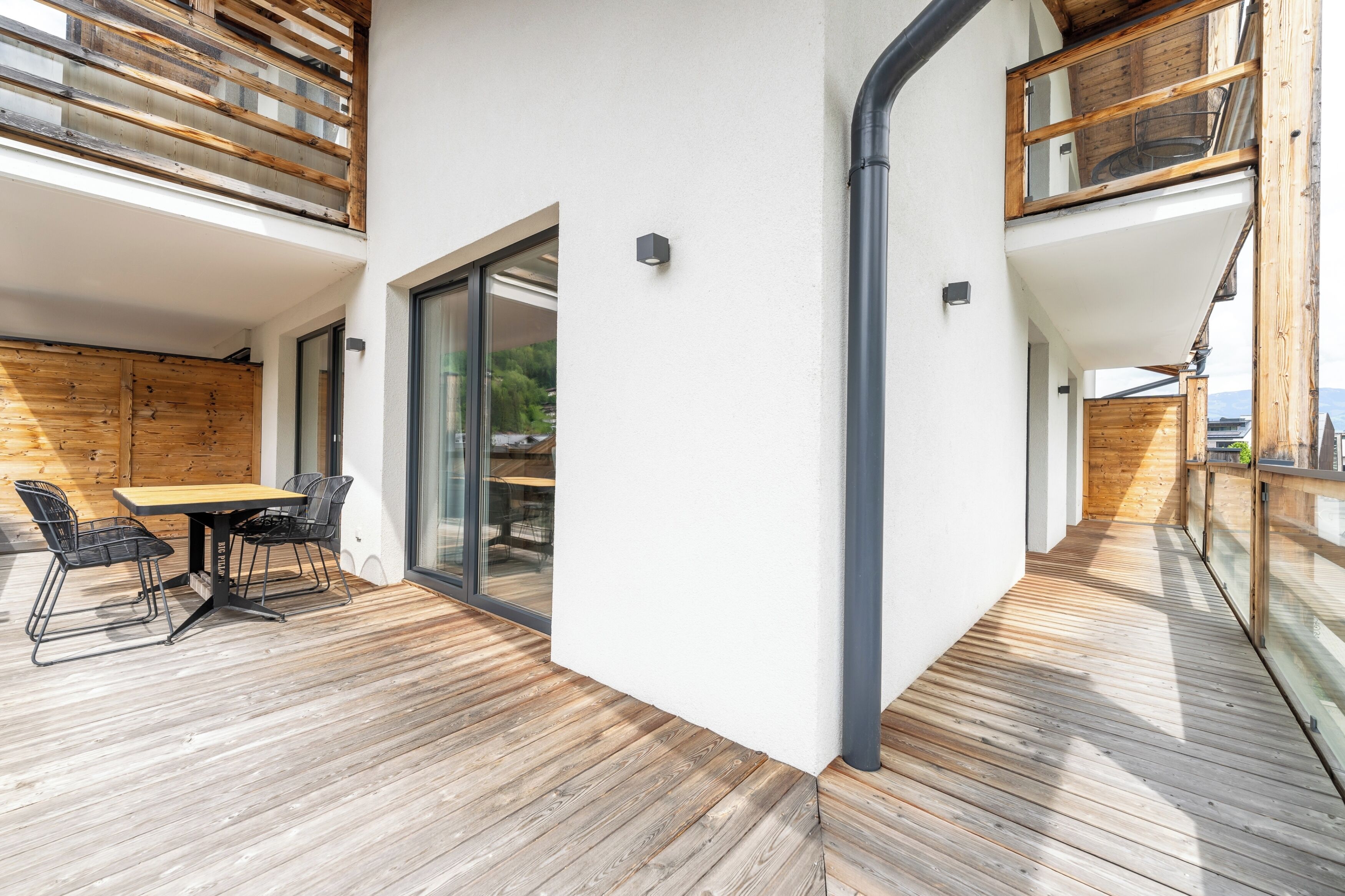 A modern outdoor dining area with a wooden table and chairs, a glass door leading to an interior space, and a wooden deck with a metal railing.