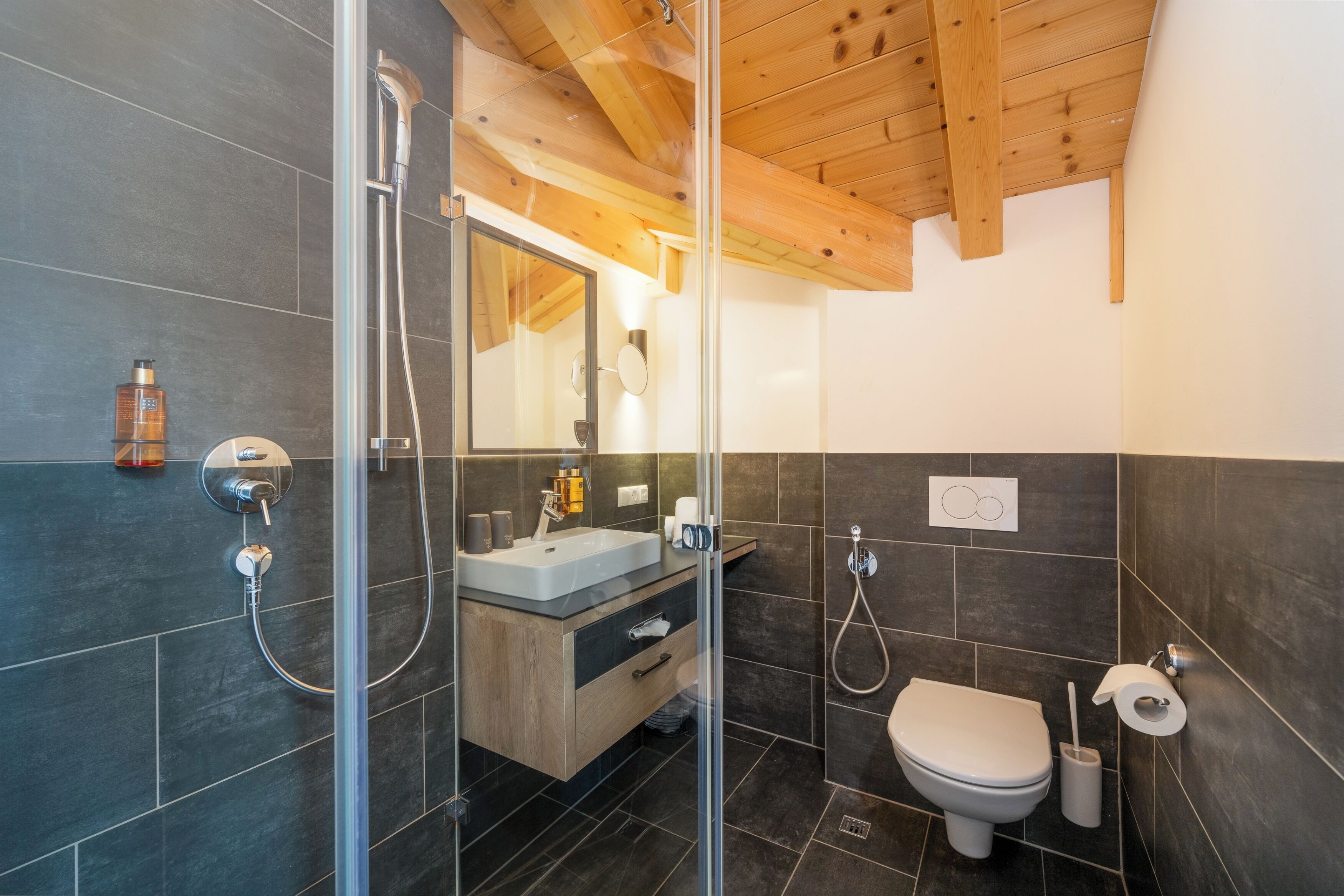 A modern bathroom with a glass shower enclosure, a wooden ceiling, and dark tiled walls.