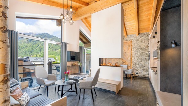 A modern interior with a stone wall, wooden ceiling beams, and a large window overlooking a mountainous landscape. - 24 by AvenidA - Mountain Hotel (Kaprun)