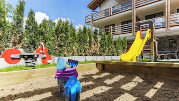 Un parque infantil con tobogán y columpios, rodeado de árboles y un edificio con balcones.