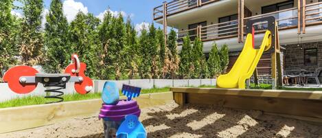 A playground with a slide and swings, surrounded by trees and a building with balconies.