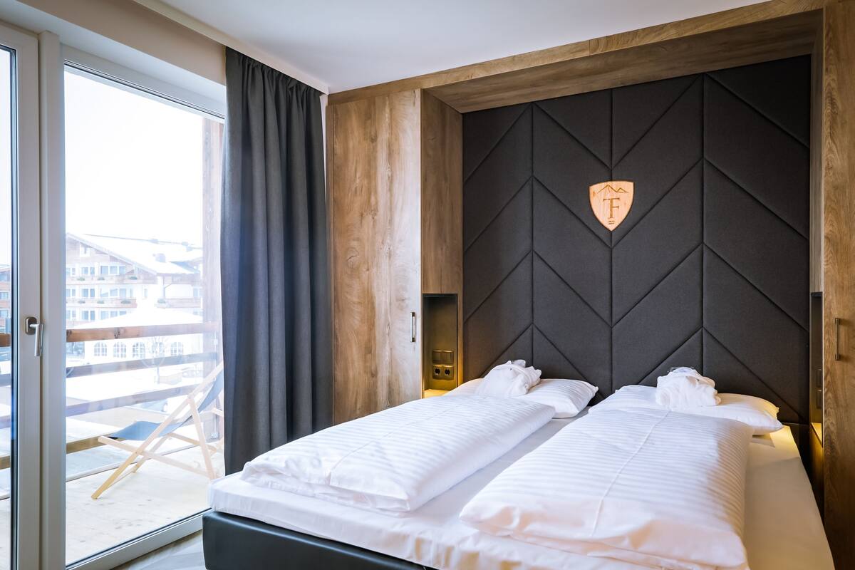A hotel room with a bed, a window with curtains, a view of buildings, and a wooden headboard.