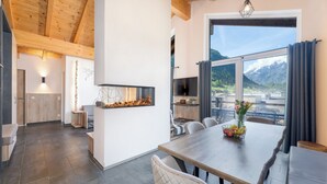 A modern dining area with a wooden table, chairs, and a view of mountains through large windows. - 24 by AvenidA - Mountain Hotel (Kaprun)