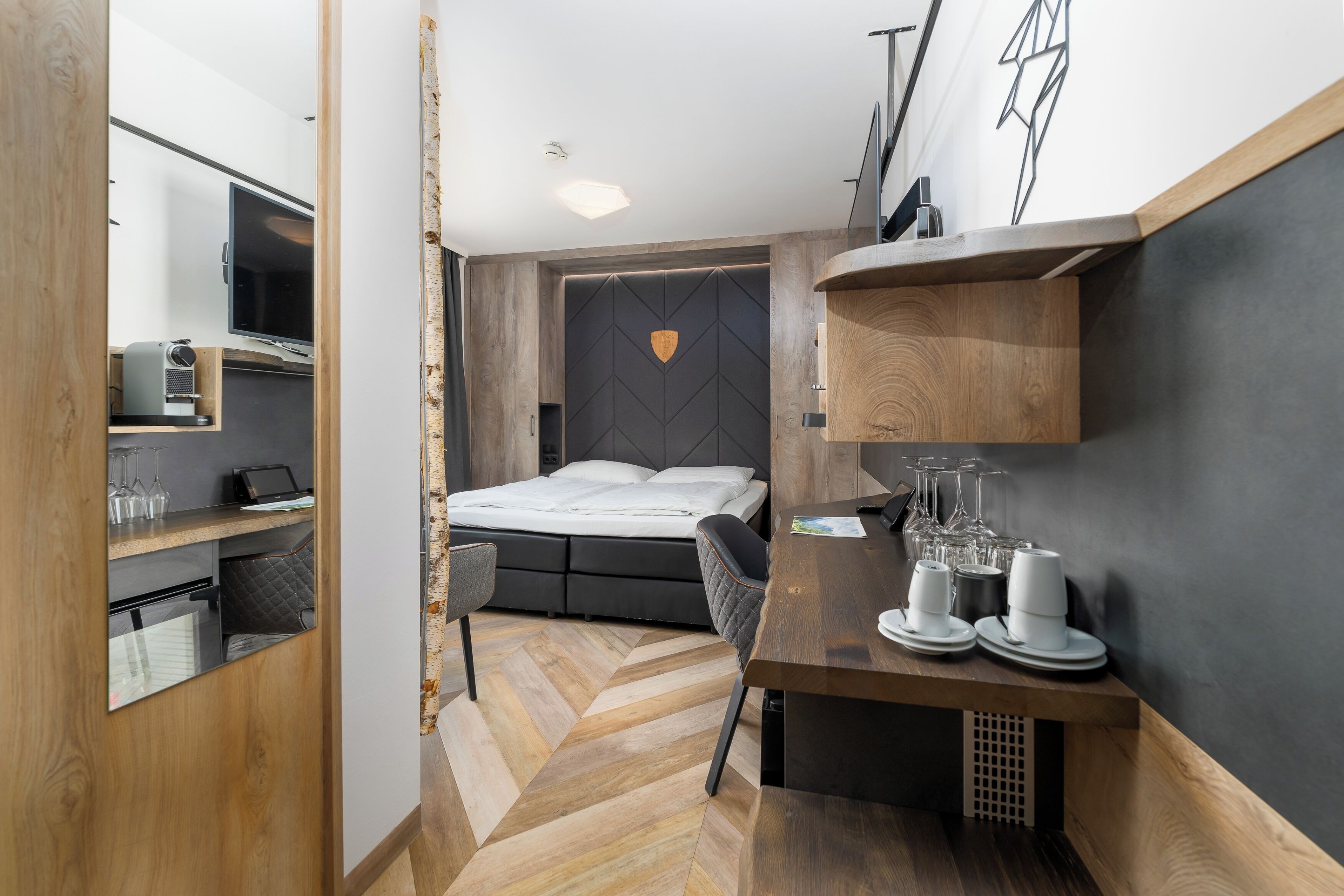 A compact hotel room with a bed, a desk with a chair, a small seating area, and a TV.