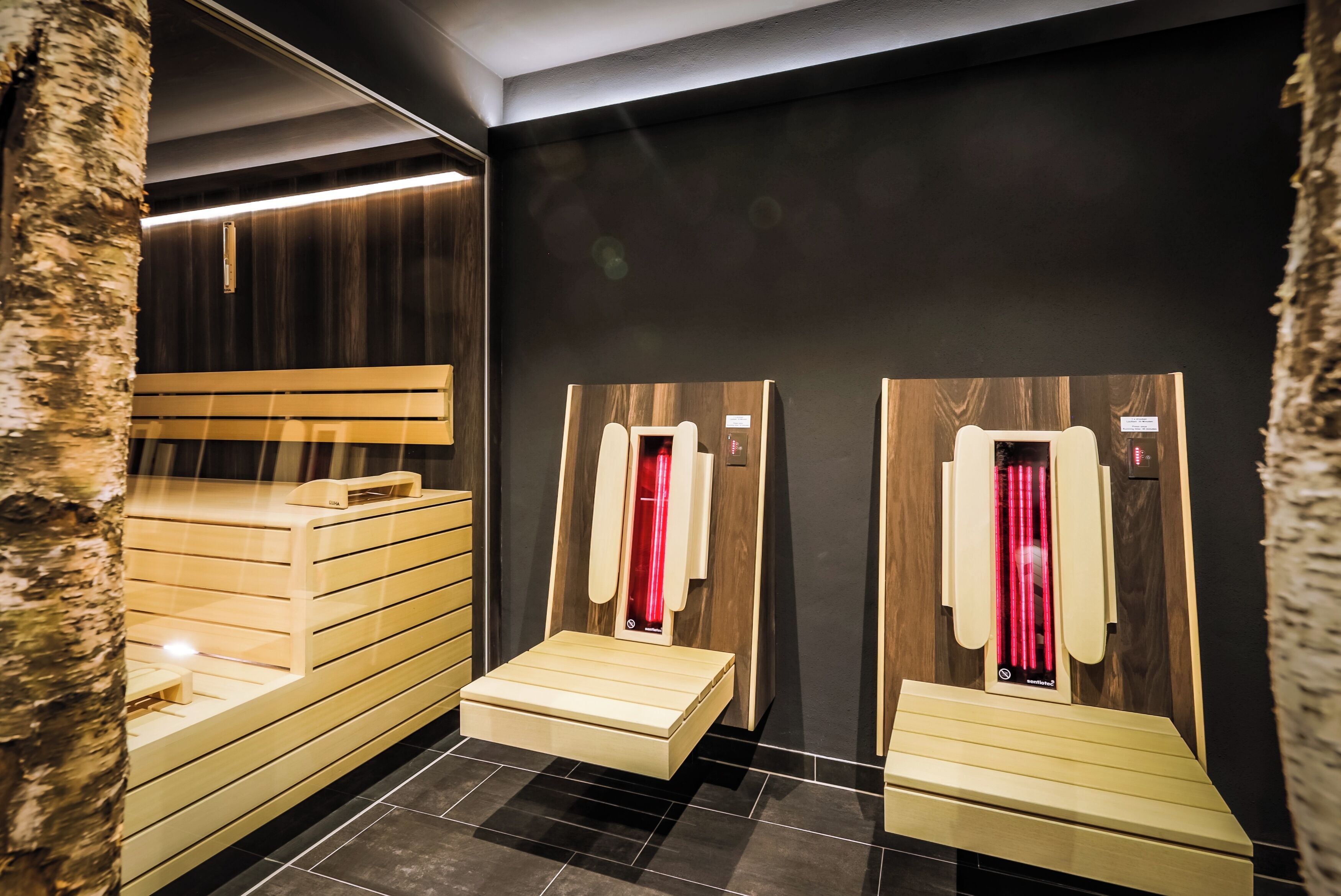 A sauna with wooden benches and steps, a wooden wall, and a black tiled floor.