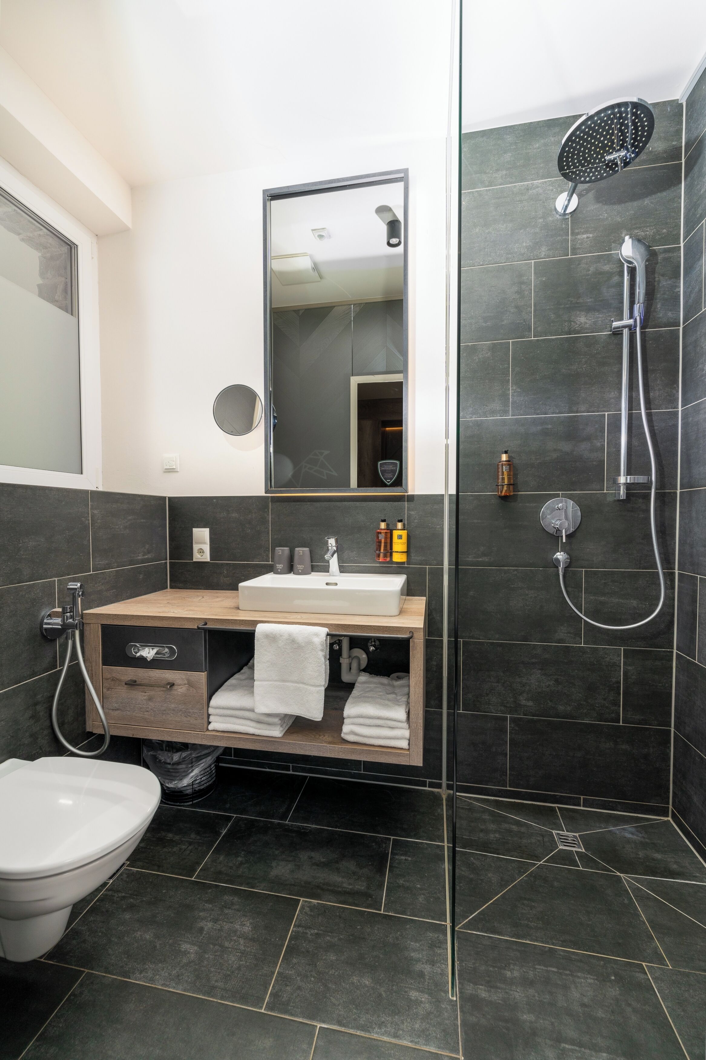 A modern bathroom with a wooden vanity, a toilet, a large mirror, and a shower area.