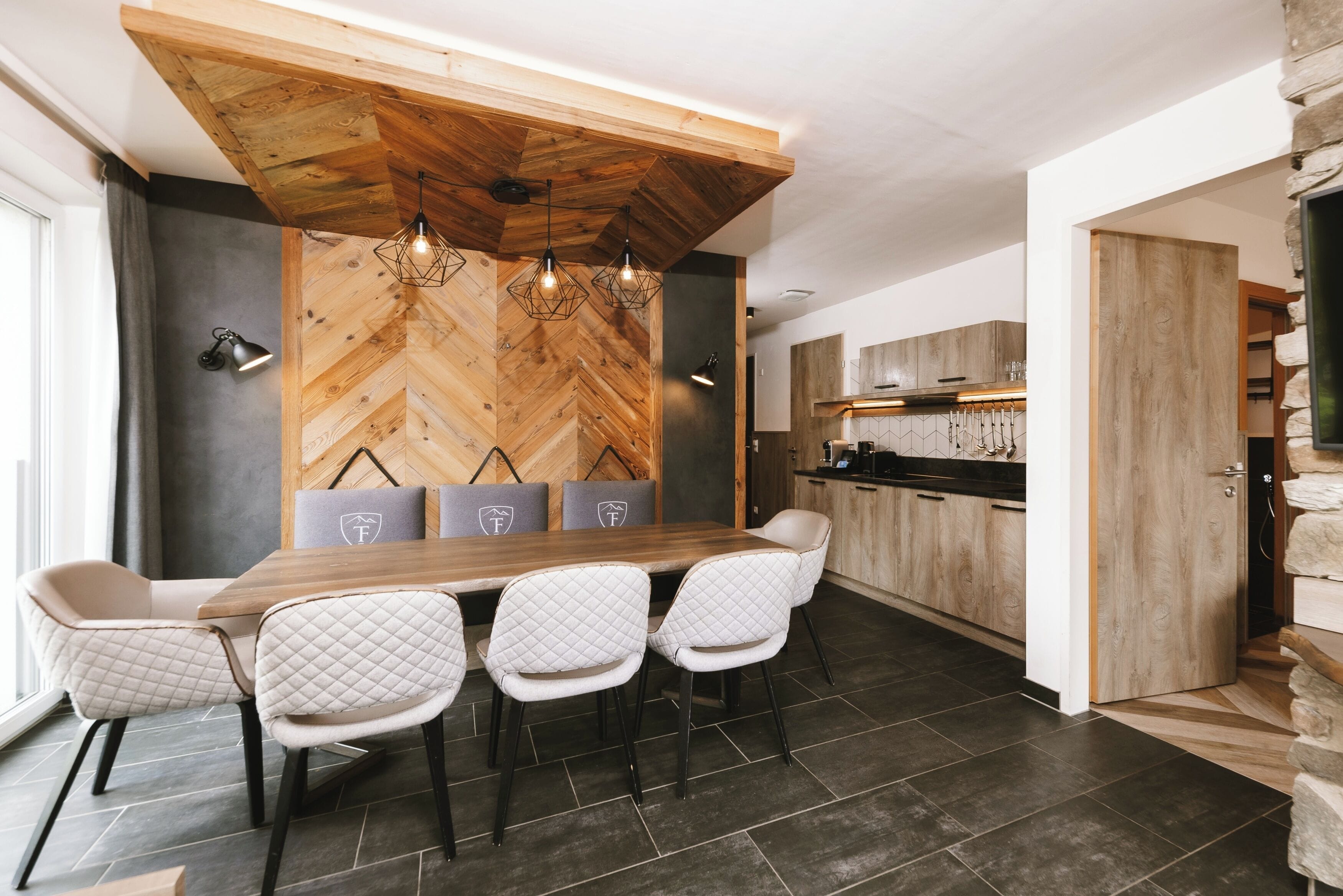 A modern dining area with a wooden herringbone wall, a long wooden table, and four quilted chairs.
