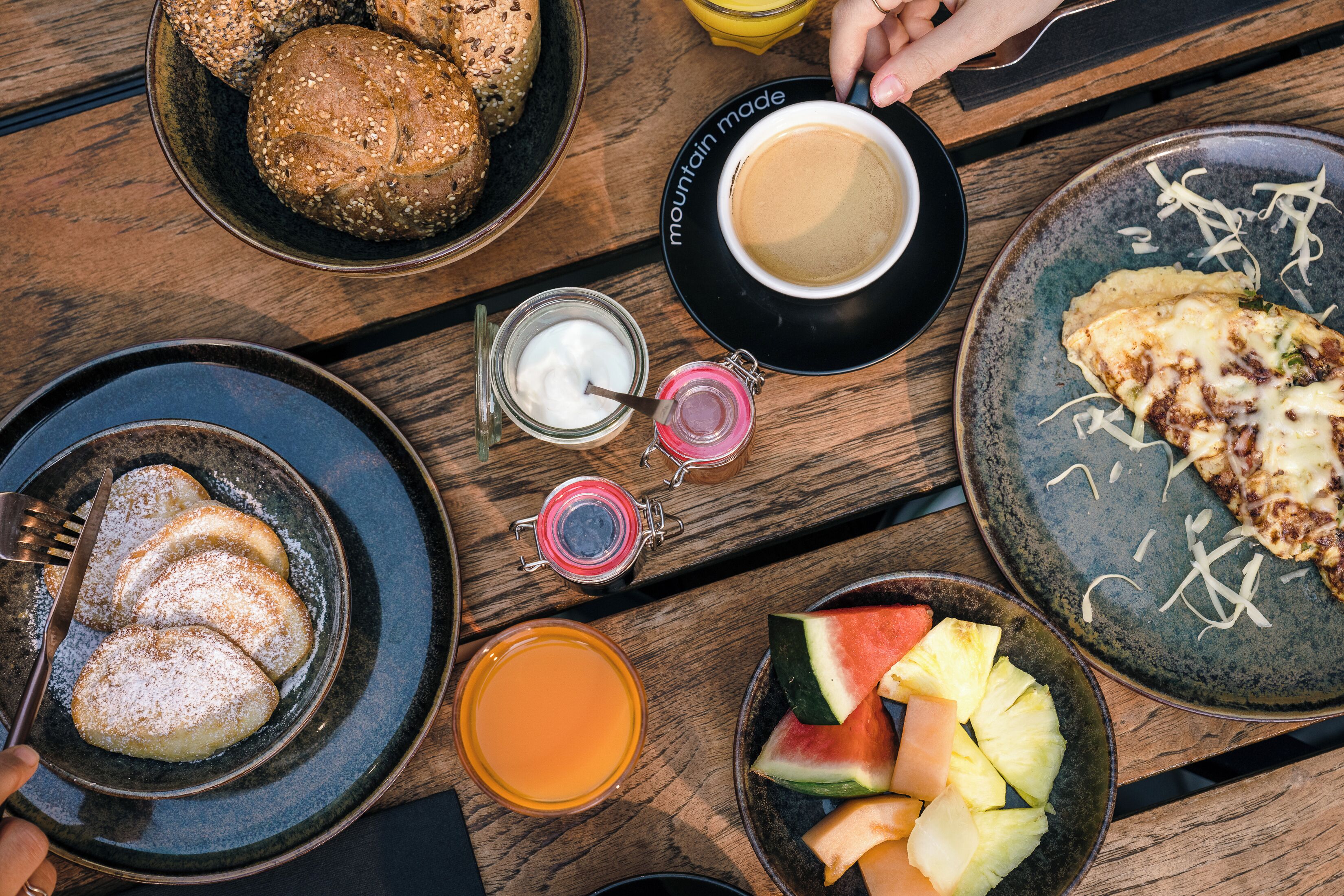 Una mesa de madera con diferentes alimentos para el desayuno, como pan, bollería, fruta y bebidas.