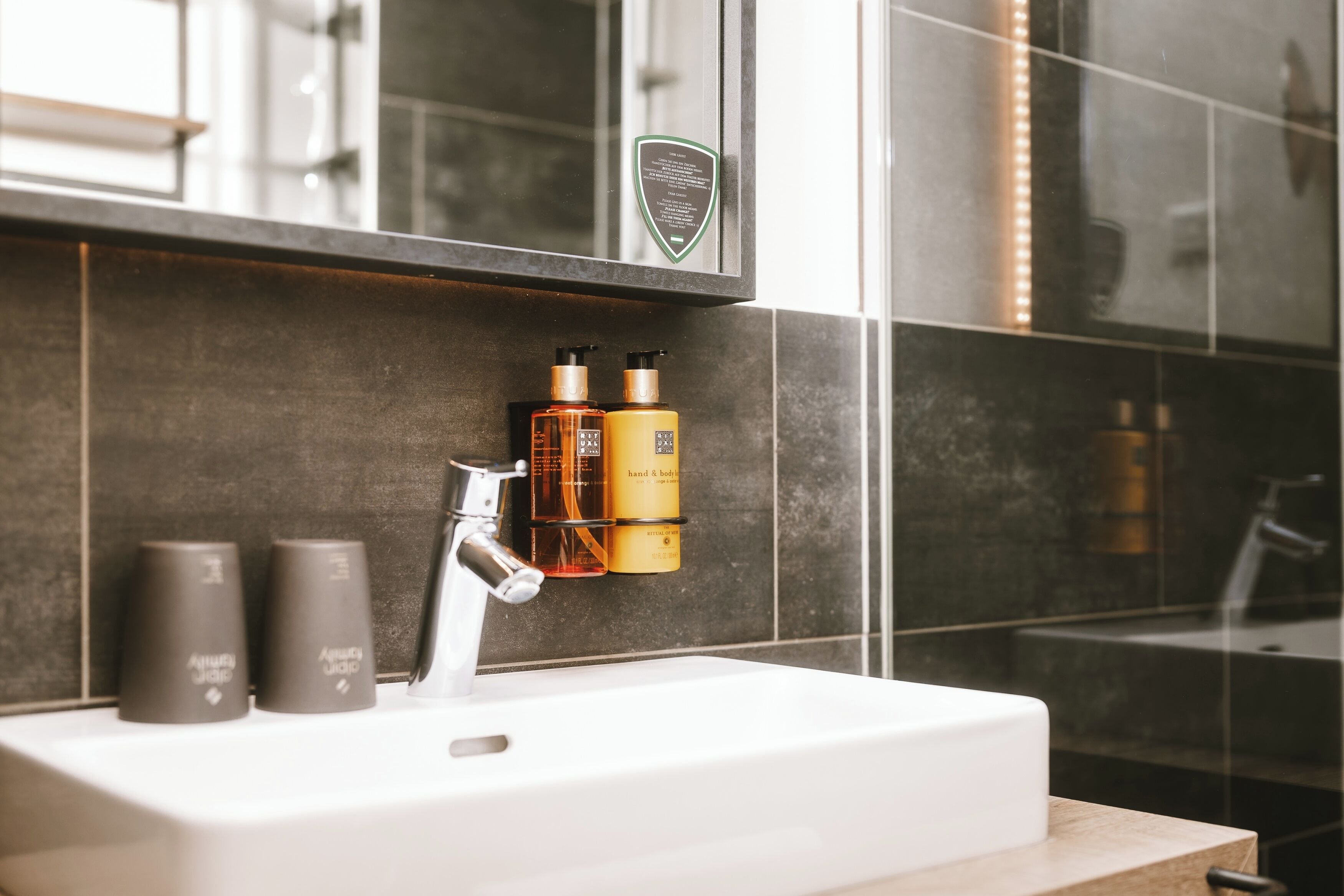A bathroom with a white sink, dark tiled walls, and a mirror above the sink. There are two bottles of hand and body wash on the wall.