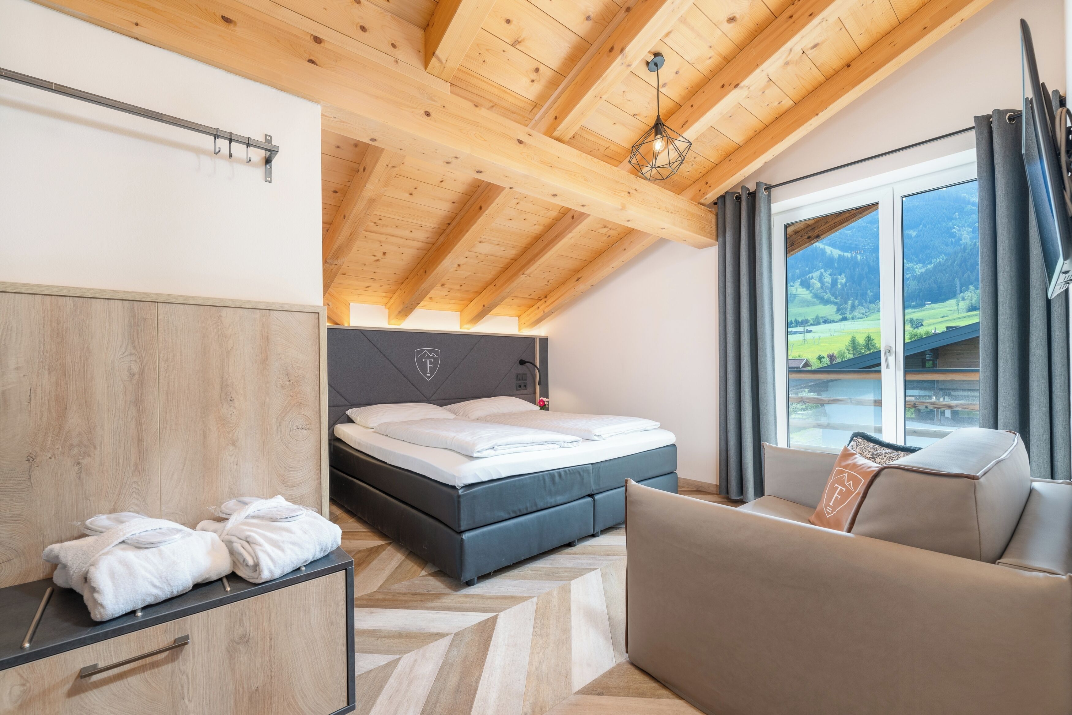 A modern bedroom with a bed, a sofa, a wooden wardrobe, and a large window with a view of a green landscape.