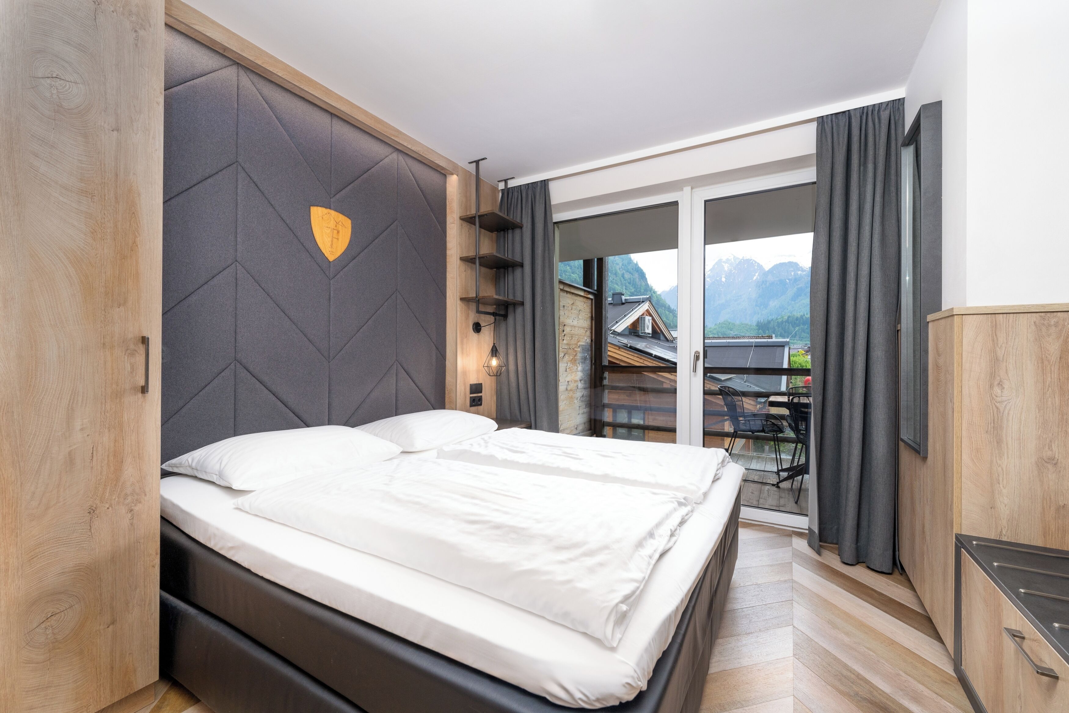 Un dormitorio moderno con cama, cabecera de madera, vistas a las montañas y un balcón con mesa y sillas.