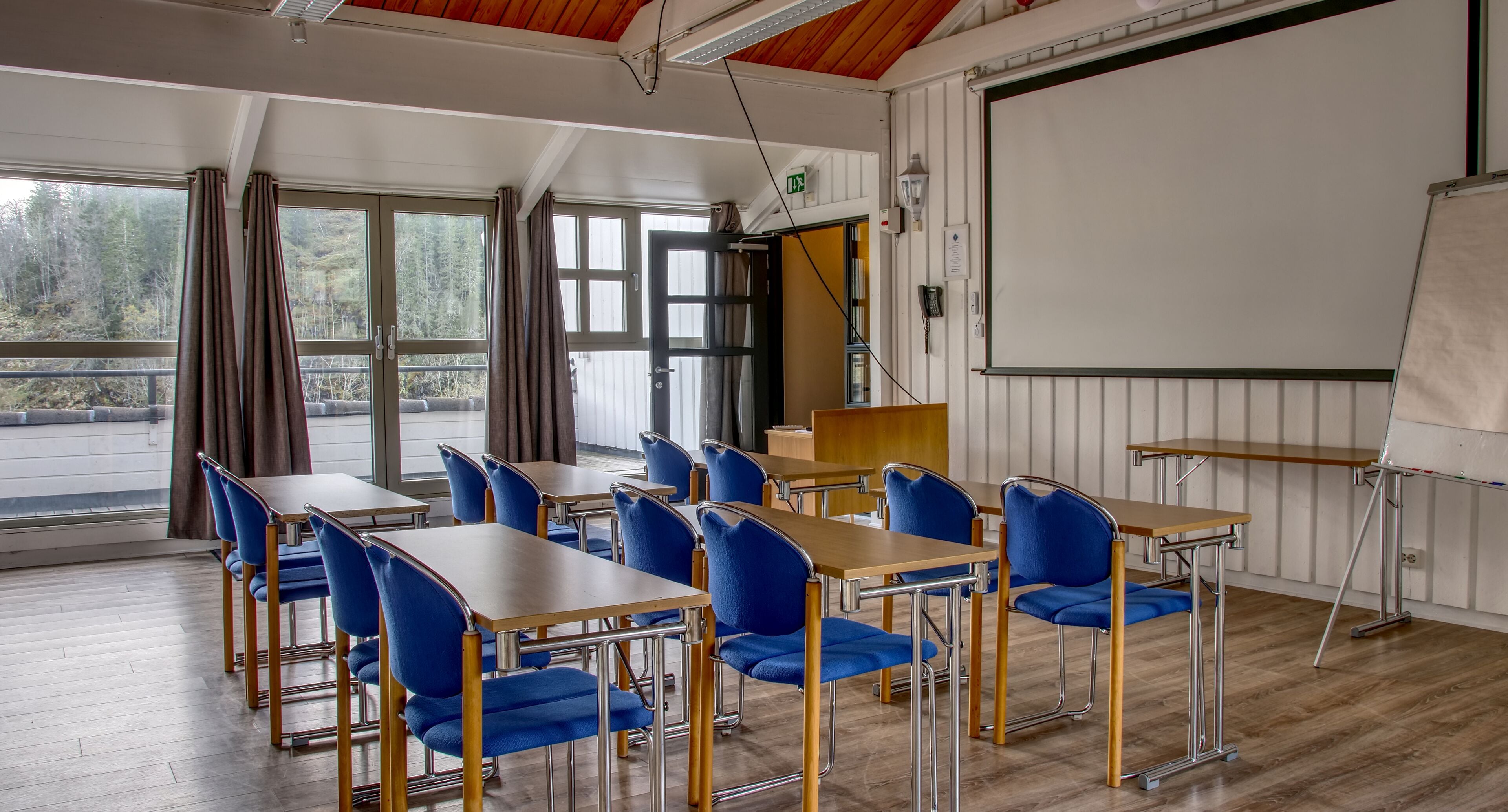 Un aula con pupitres de madera y sillas azules, ventanas amplias y una pantalla para proyector.