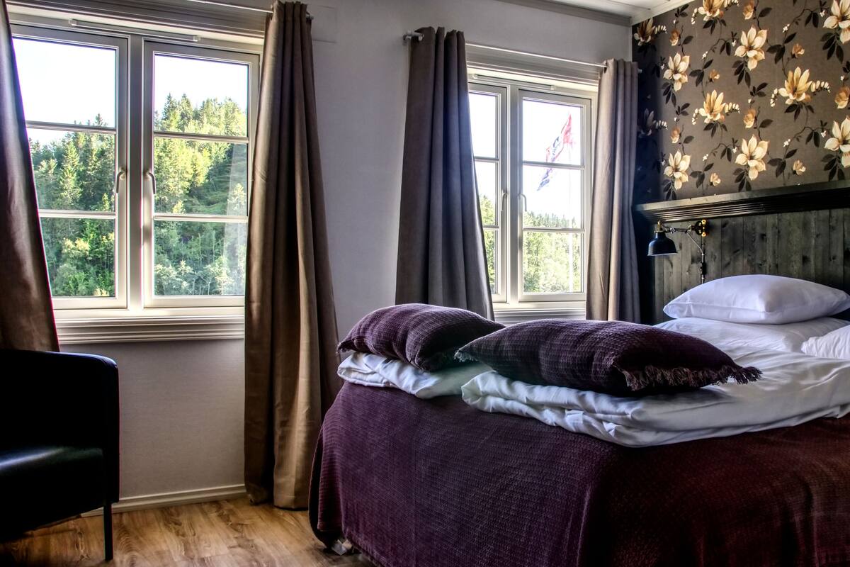 Een slaapkamer met een bed, gordijnen, een raam met uitzicht op bomen en bloemenbehang.