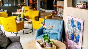 Een moderne hotel lobby met gele en blauwe stoelen, een ronde tafel met een bloemenvaas, en een schilderij aan de muur.