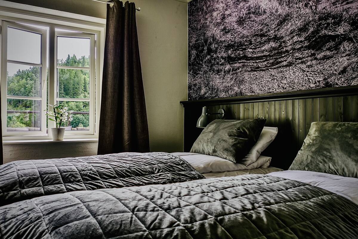 Een slaapkamer met twee bedden, een raam met gordijnen, een bedlampje en een groot wandtapijt.