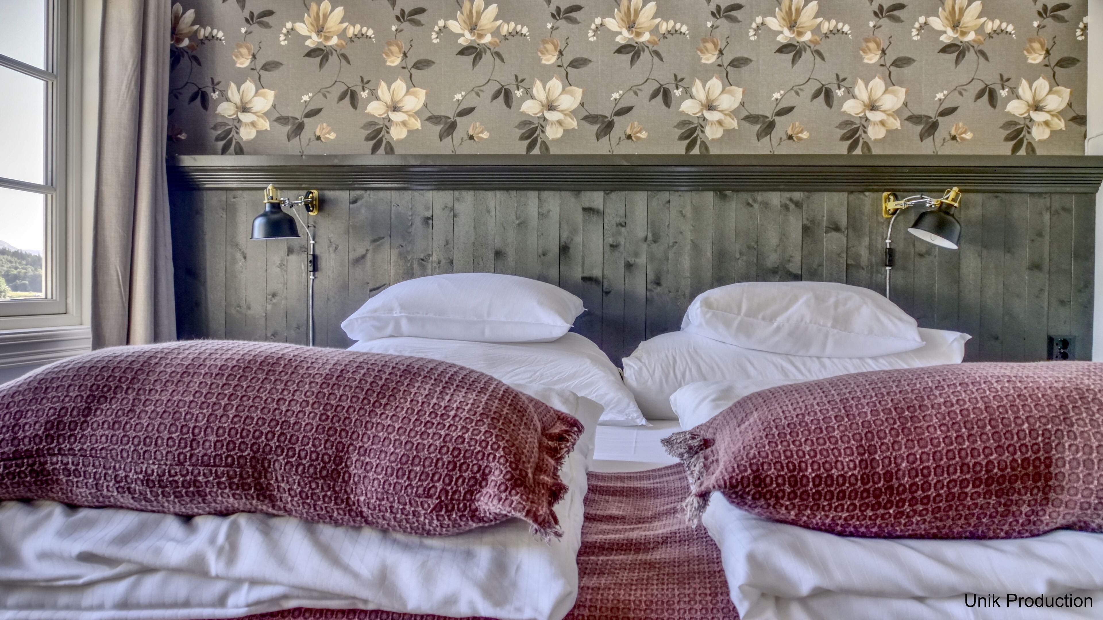 Una habitación con dos camas, cabecero de madera, papel tapiz floral y una ventana con cortinas.