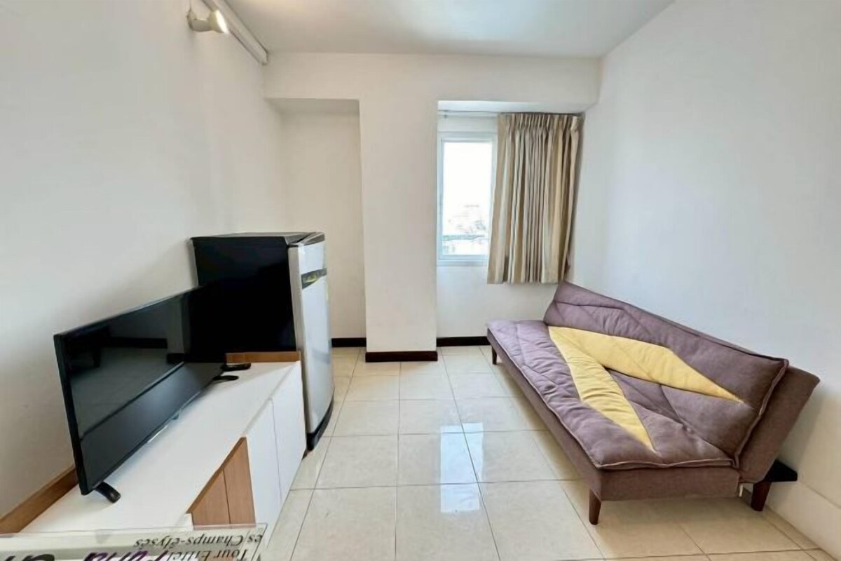 Una sala de estar con televisión de pantalla plana, un sofá, una ventana con cortinas y piso de baldosas.