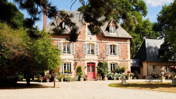 A two-story house with a red door, multiple windows with shutters, and a garden with potted plants and a fountain. - Manoir des Lions de Tourgéville (Tourgeville)