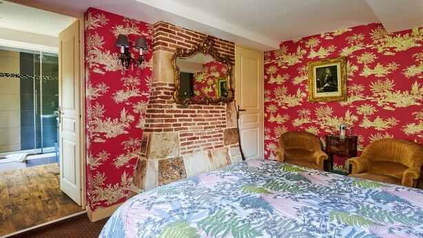A bedroom with a brick wall, a large bed with a floral bedspread, two chairs, a mirror, and a picture frame on the wall. - Manoir des Lions de Tourgéville (Tourgeville)
