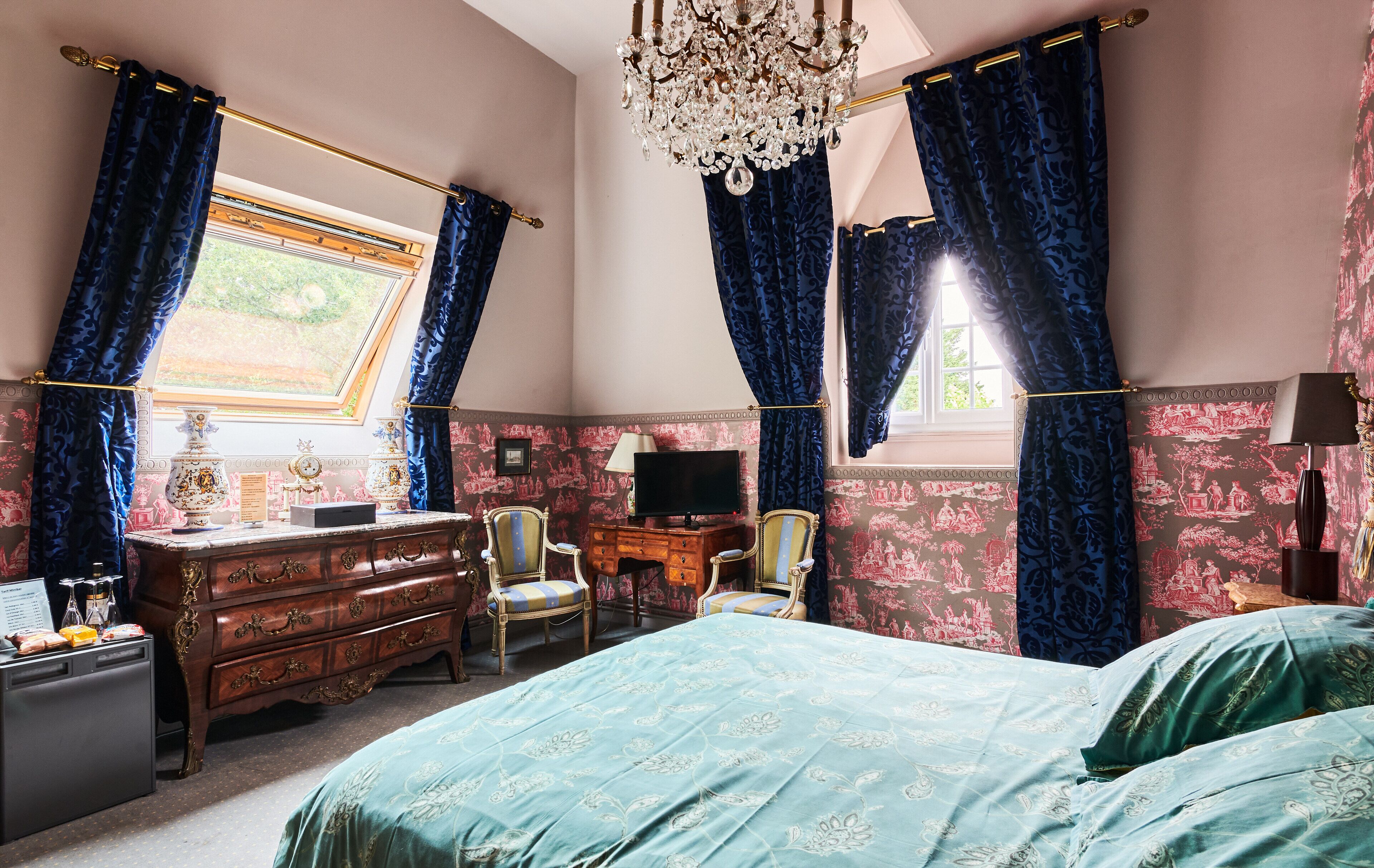 A bedroom with a large bed, a dresser, two chairs, a desk with a television, and a chandelier.