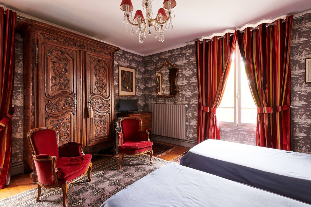 Ein Zimmer mit einem großen Holzschrank, zwei roten Sesseln, einem Bett und einer Kandelaber.