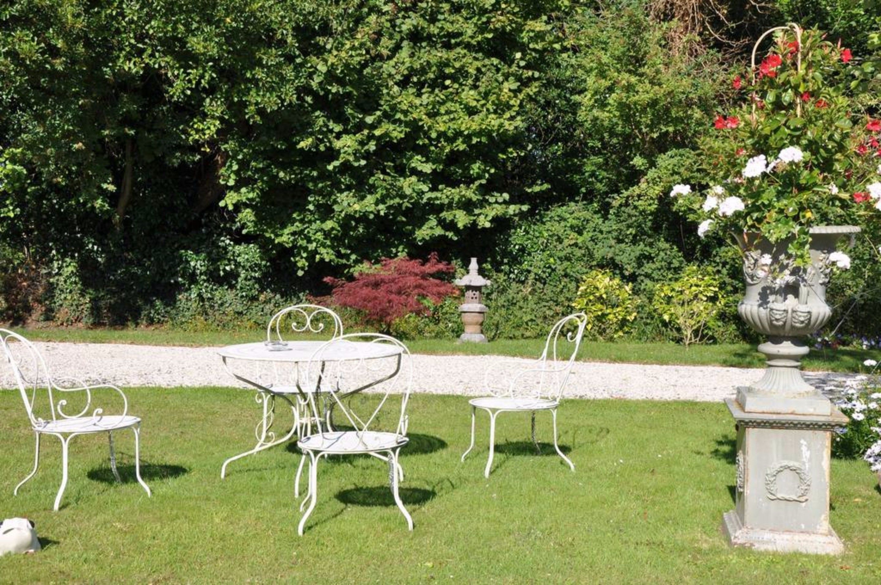 A garden with a white metal table and chairs, surrounded by lush greenery and a decorative urn.