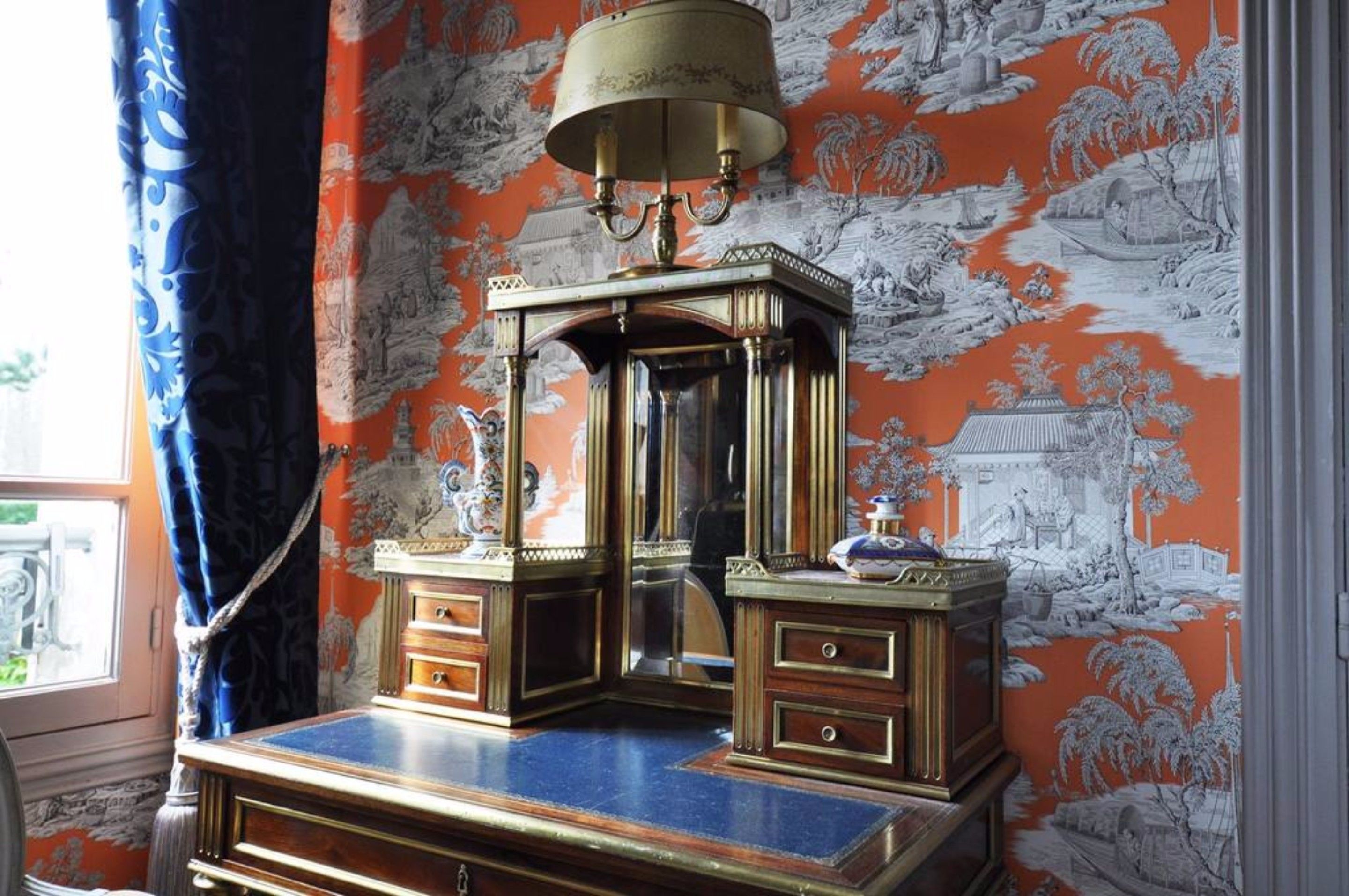 Ein Zimmer mit einem traditionellen Holzschrank, Spiegel, einer Messinglampe und einem Wandpapier mit Muster.