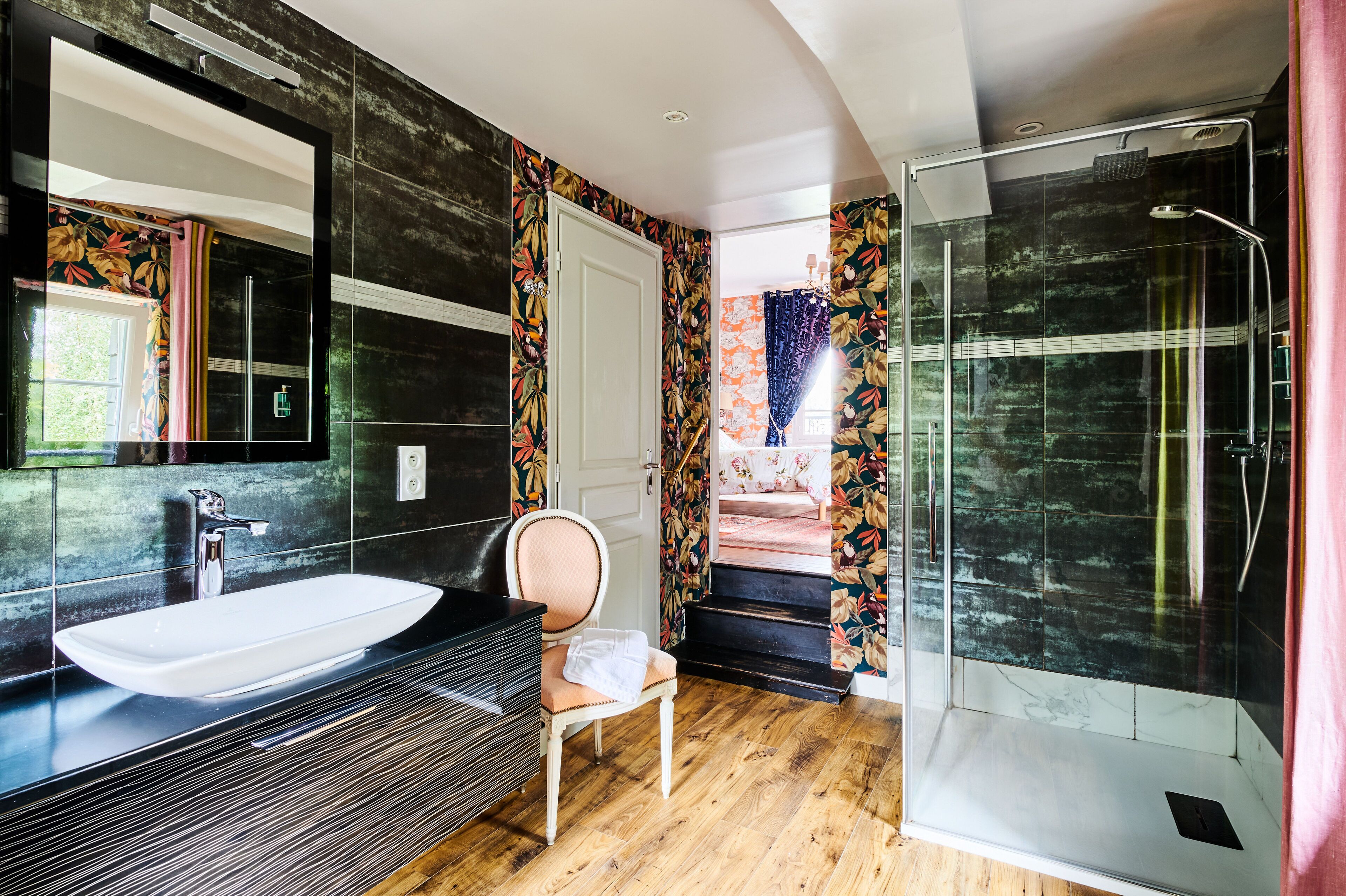 A modern bathroom with a large mirror, a white sink, a shower, and a chair.