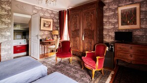 A hotel room with a bed, two red armchairs, a wooden wardrobe, a desk with a chair, a television, and a chandelier. - Manoir des Lions de Tourgéville (Tourgeville)